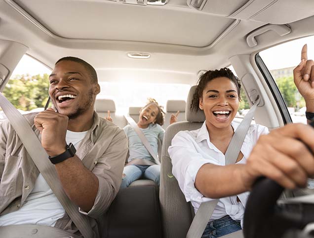 A family is smiling brightly while gesturing and singing along to a song in their car.