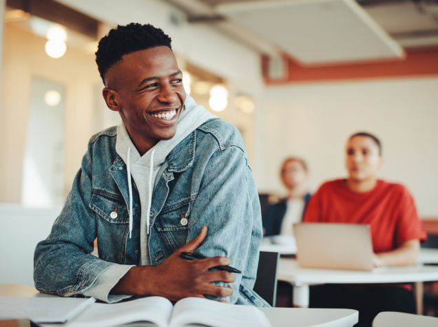 A young man in a classroom is smiling off to his side at something out of the frame.