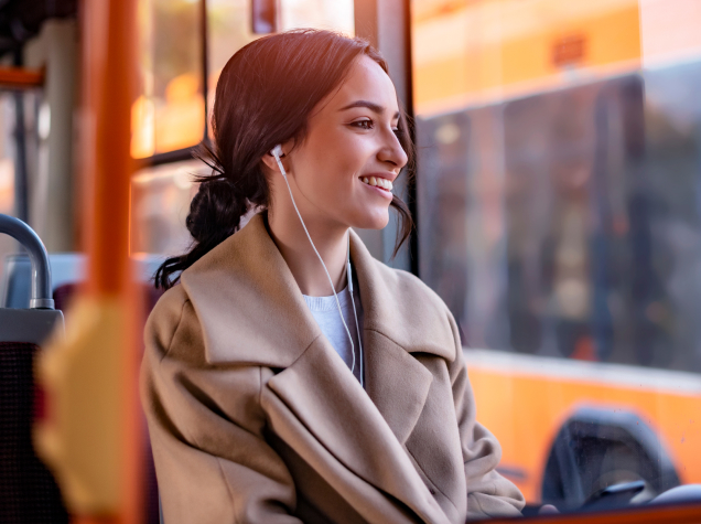 A young woman is sitting in a bus and staring out her window while listening to music through her earphones