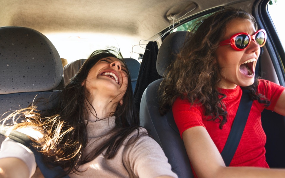 Two teenage girls in a car. The one on the left is laughing and the one on the right is smiling and looking ahead.