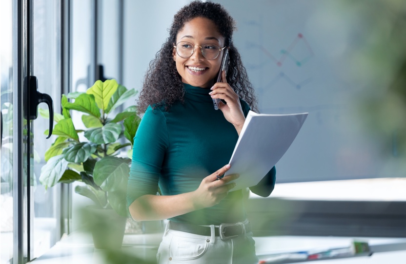 A young woman is holding paperwork while on the phone.