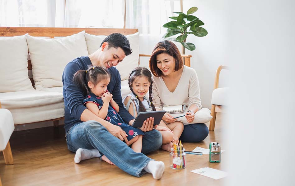 A couple and their two young girls sit in front of a couch watching something together on a tablet. 