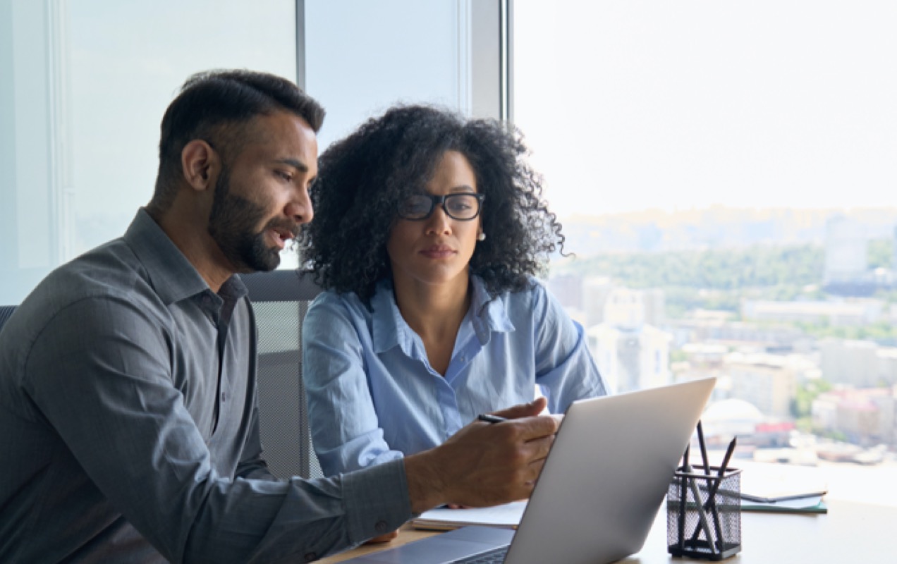 A man and a woman, sitting side-by-side in an office overlooking a city, looking at a laptop.
