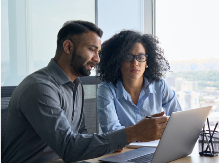 A man and a woman, sitting side-by-side in an office overlooking a city, looking at a laptop.