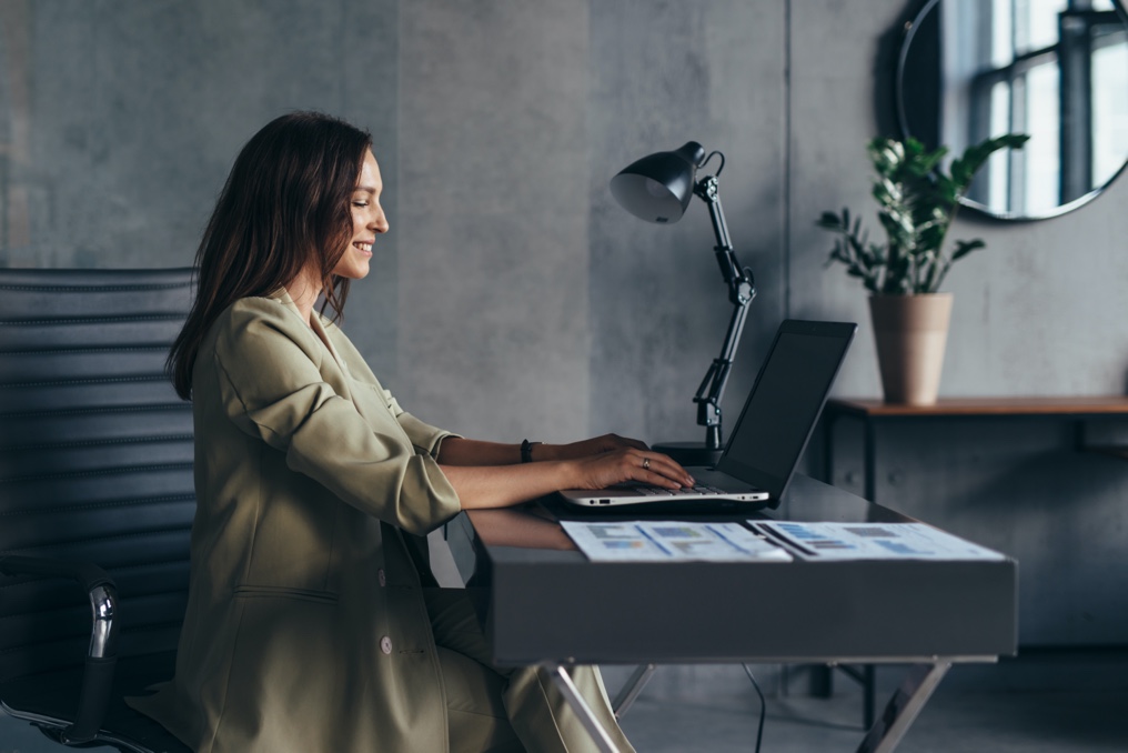 Smiling woman sits at her desk in a gray office, typing on laptop.