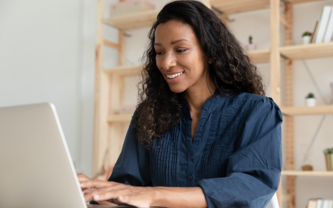 Smiling woman in a blue shirt focuses on the resources on her laptop, as she researches and learns how to invest.