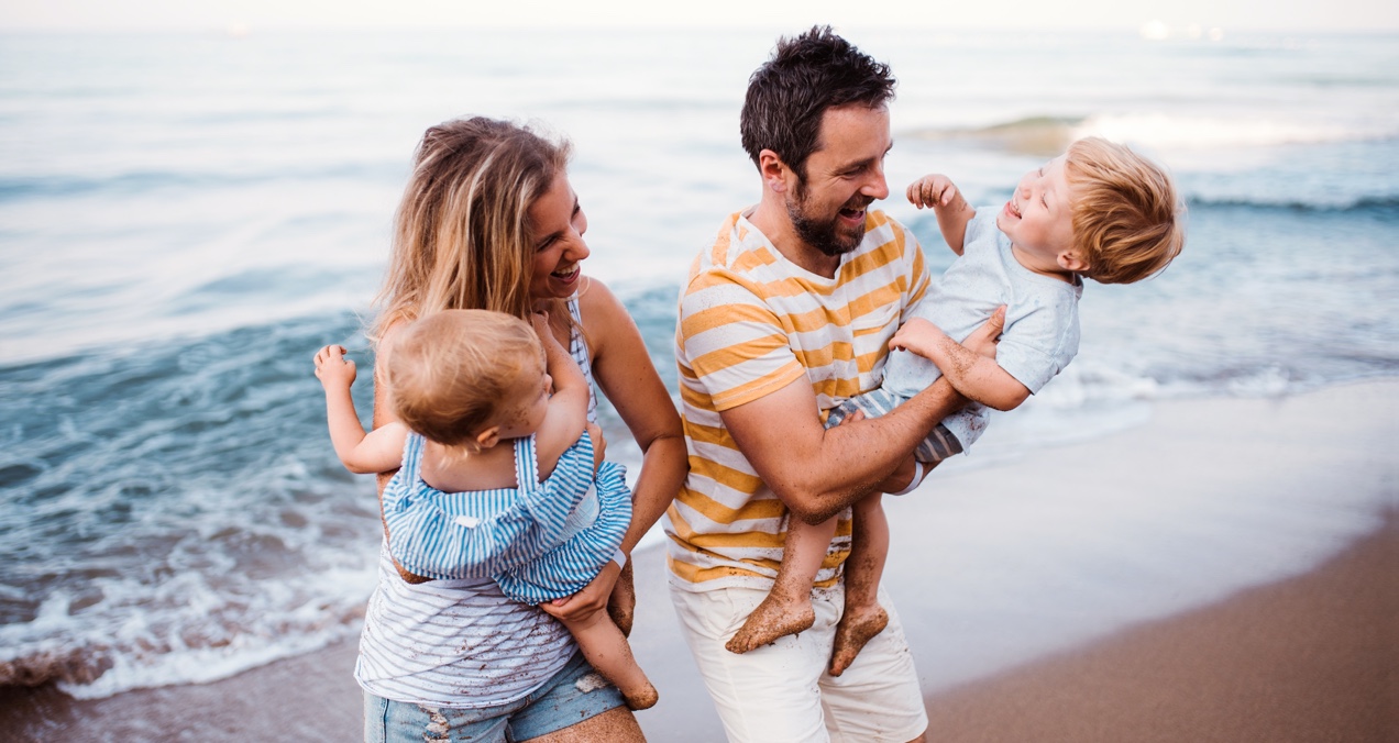 Laughing family of four in matching striped outfits plays on the beach with waves behind them. Each parent holds a child.