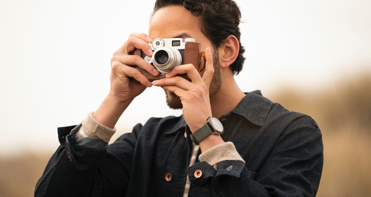 Wavy-haired man in a denim jacket holds a camera, encased in leather, in front of his face as he focuses on taking a picture.