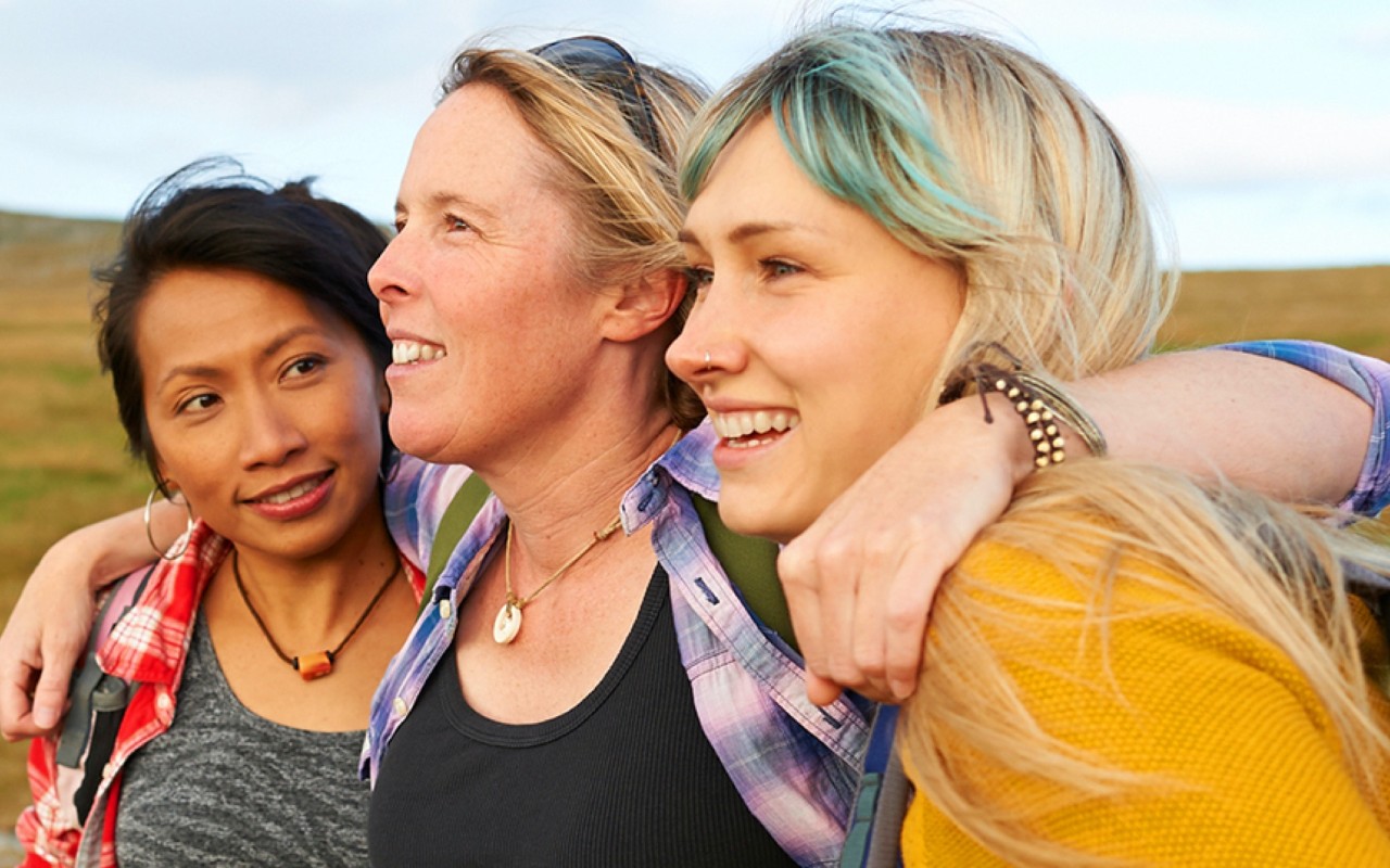 Three smiling women stand in a field with their arms around each other. 