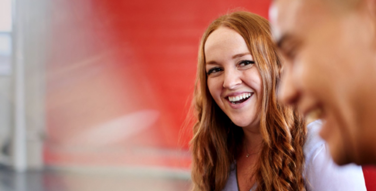Laughing woman looks toward a man in the foreground in front of a red background.