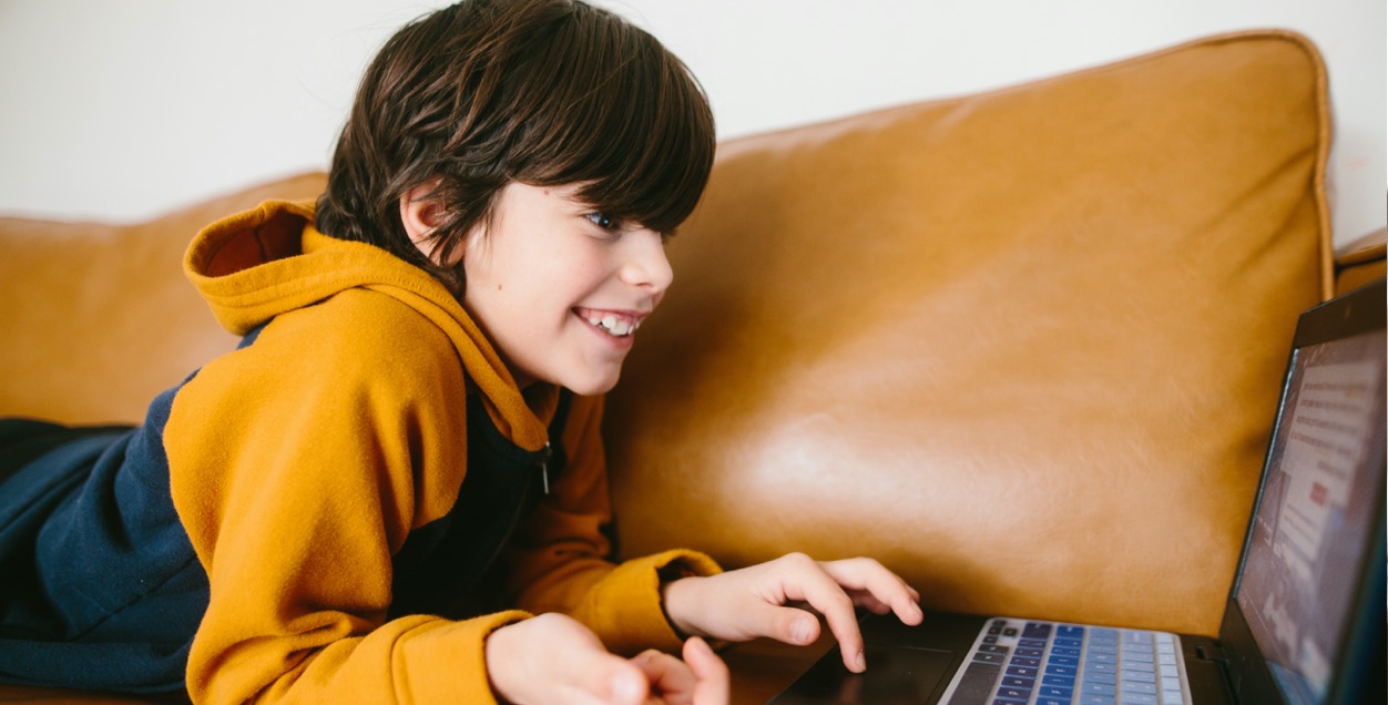 Grinning little boy looks at a laptop as he lies on his stomach on a couch.