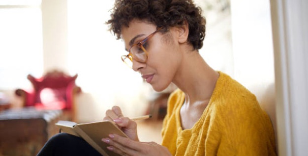 Woman in glasses looks intently at a journal as she writes, sitting in a light-filled room.