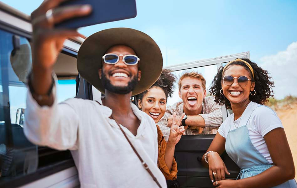 A group of four friends are standing next to a vehicle. The person in front is taking a selfie and smiling while wearing a white shirt, a hat, and sunglasses. The three others are looking at the camera with smiles and wearing casual clothes.