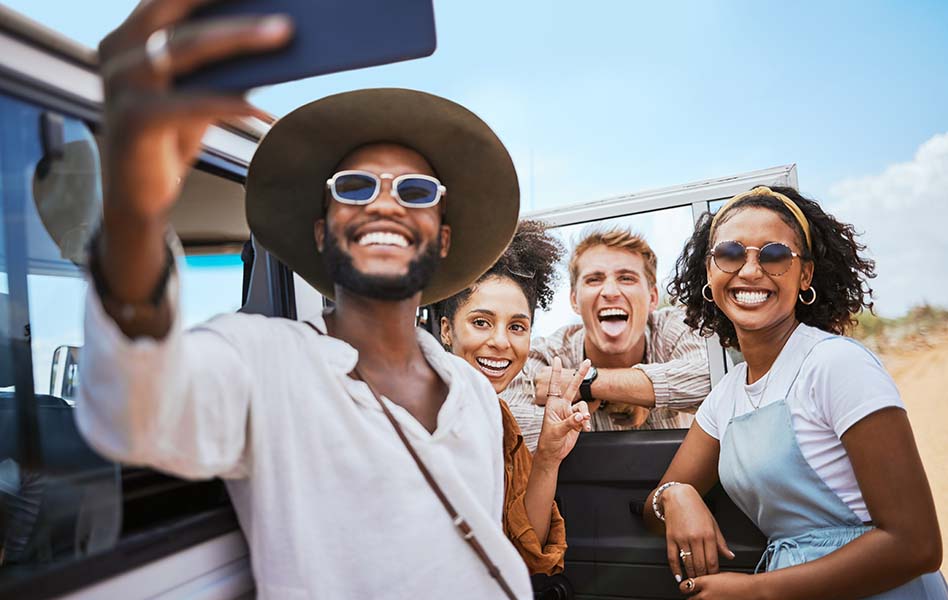 A man takes a selfie with three of his friends standing in front of a jeep on the beach. 