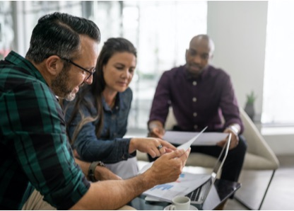 Group of two men and a woman sit in an office together, discussing their taxes with paper documents and a laptop. 