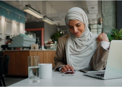 Smiling woman in a hijab is sitting in a cafe with her laptop, looking down at her phone as she calculates her tax rate. 