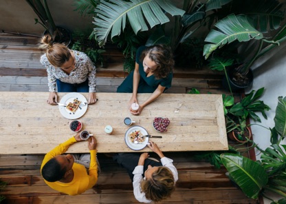 Camera looks down on a plant-filled room with a wood table, where four women sit with their breakfasts in front of them. 