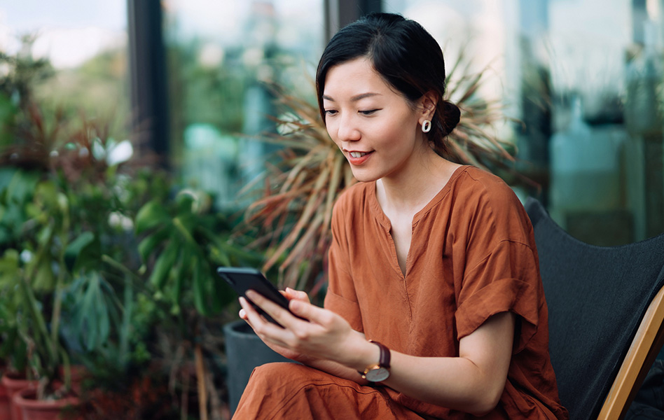A woman in an orange shirt sitting in front of a plant checks her phone.