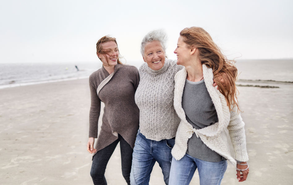 Older woman walking arm-in-arm with her two adult daughters on the beach.