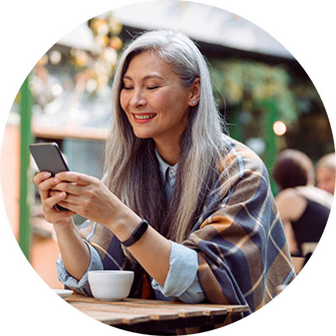 A woman sitting, drinking coffee, and smiling at her phone.