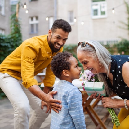  Person stands smiling with their hand on the back of a child who looks up and smiles at another person who is smiling and leaning toward the child so their faces almost touch.