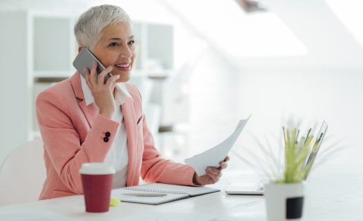 Person sits smiling at a desk with a laptop on it while on the phone and holding papers in their other hand.