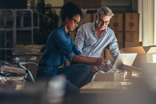 Two smiling people sit at a work bench in a supply room and look at an open box.
