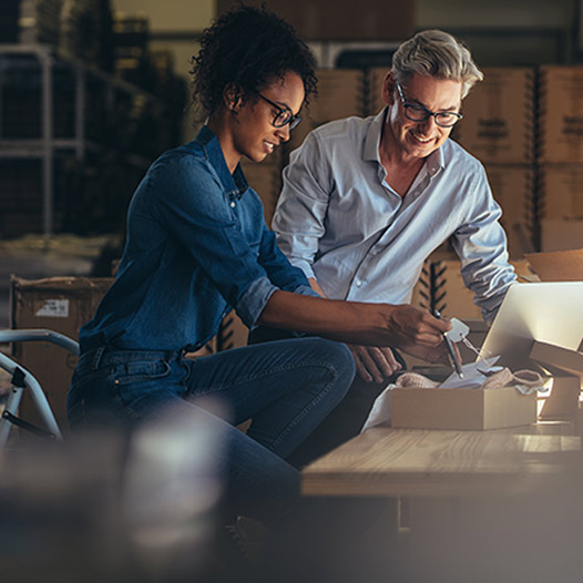 Two smiling people sit at a work bench in a supply room and look at an open box.