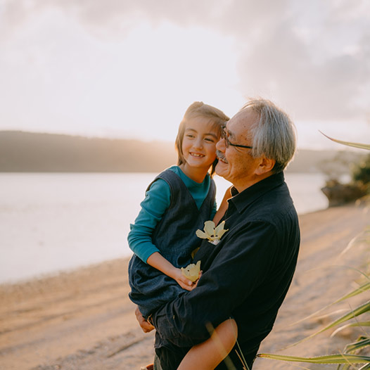 Person smiles and holds a child while standing outside near a body of water with mountains in the background.