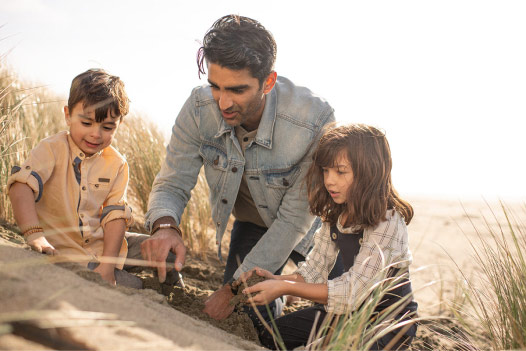 Person and two children dig in the sand near a body of water.