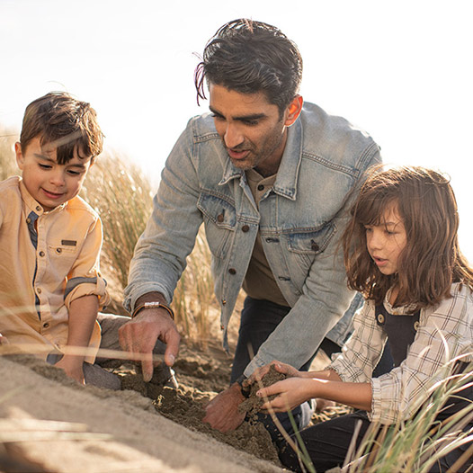 Person and two children dig in the sand near a body of water.