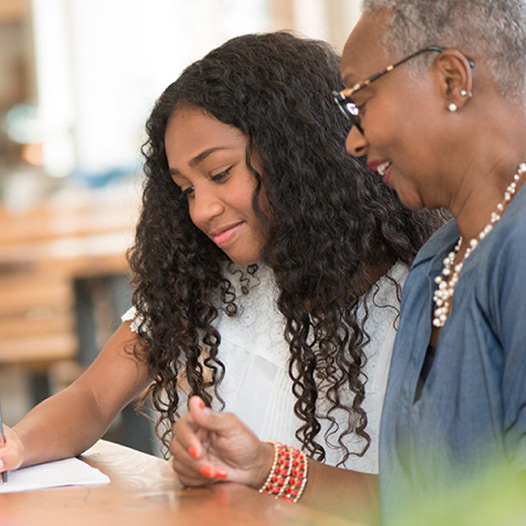 Child holds a pen and writes on a paper while a person stands nearby and looks down at the paper .