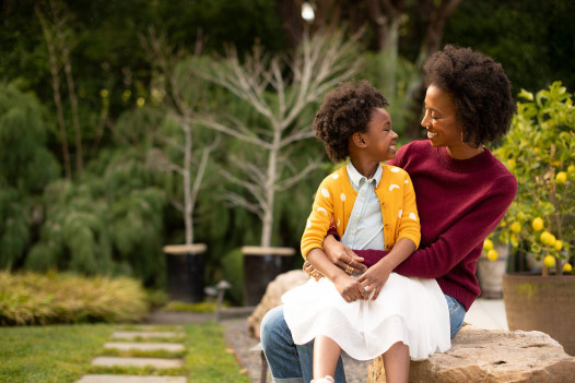 Child sits on a person’s lap and they smile at each other while outside in a garden.