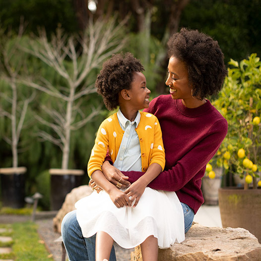 Child sits on a person’s lap and they smile at each other while outside in a garden.