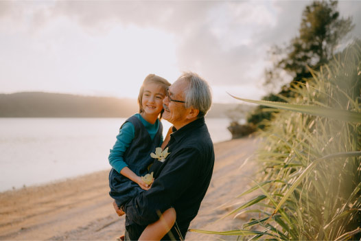 Person smiles and holds a child while standing outside near a body of water with mountains in the background.