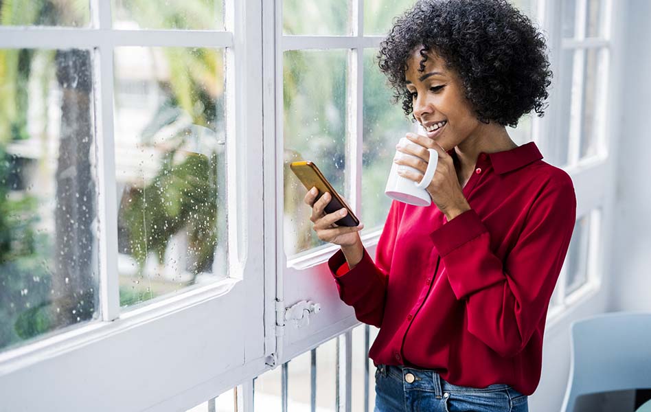 A young woman standing by the window is holding a mug while looking at her smartphone.